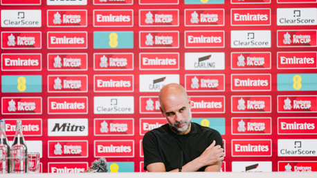 A person seated at a press conference table in front of a wall covered with FA Cup and sponsor logos. Bottled water is visible on the table.