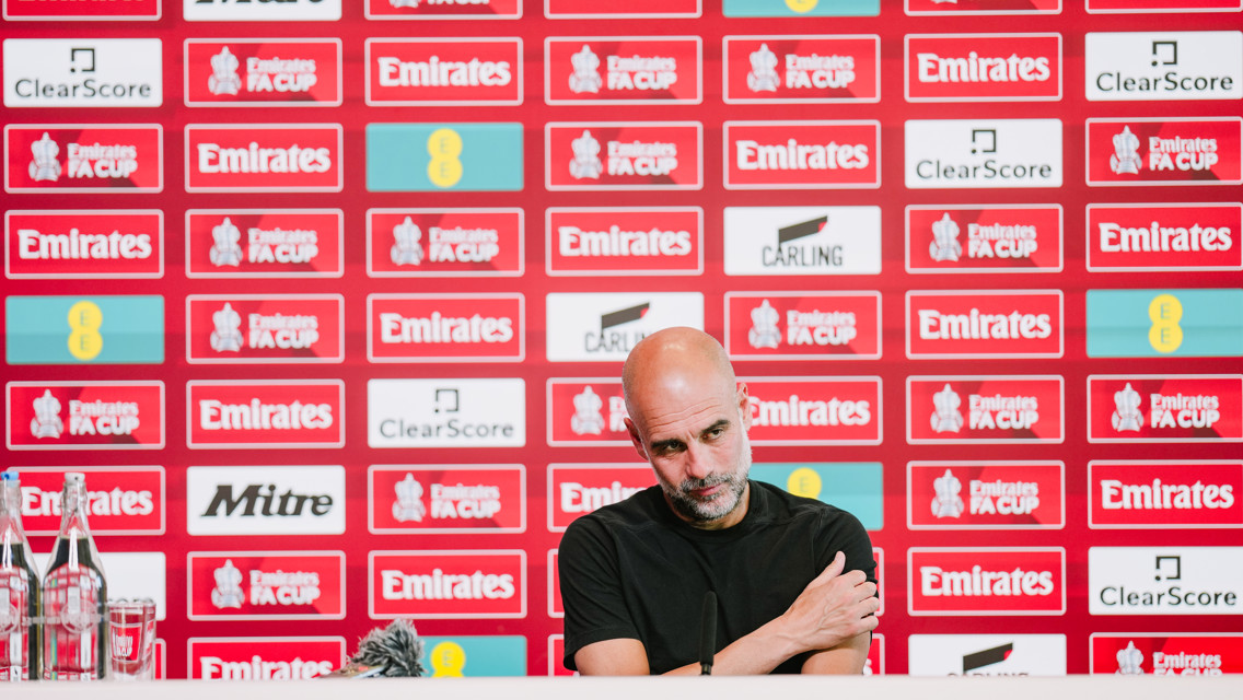 A person seated at a press conference table in front of a wall covered with FA Cup and sponsor logos. Bottled water is visible on the table.