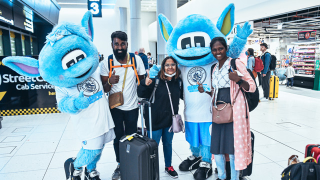 A group of travelers with luggage pose for a photo with two Manchester City mascots, Moonchester and Moonbeam, at an airport.