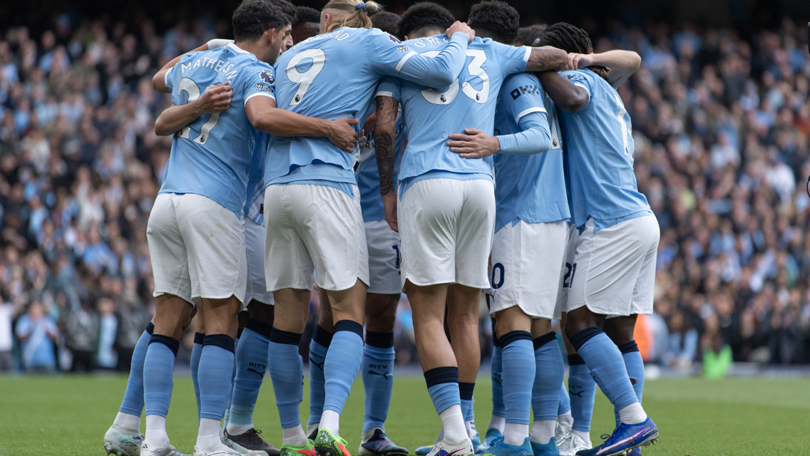 The image depicts Manchester City players in a team huddle wearing their light blue home kit during a match.