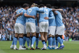 The image depicts Manchester City players in a team huddle wearing their light blue home kit during a match.