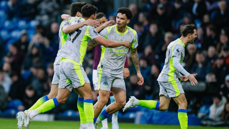 Soccer players wearing grey and neon kits celebrate a goal with fans in the background.
