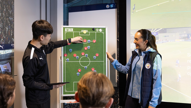Two coaches in Manchester City attire demonstrate tactics using a magnetic football board to an audience.