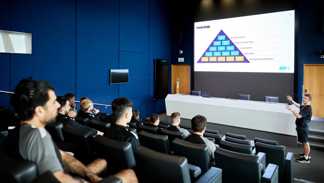 A group of young people watching a presentation in a conference room. The presentation slide appears to show a pyramid chart labeled 'Practice Design' and is branded with the Manchester City Electric Blue Logo.