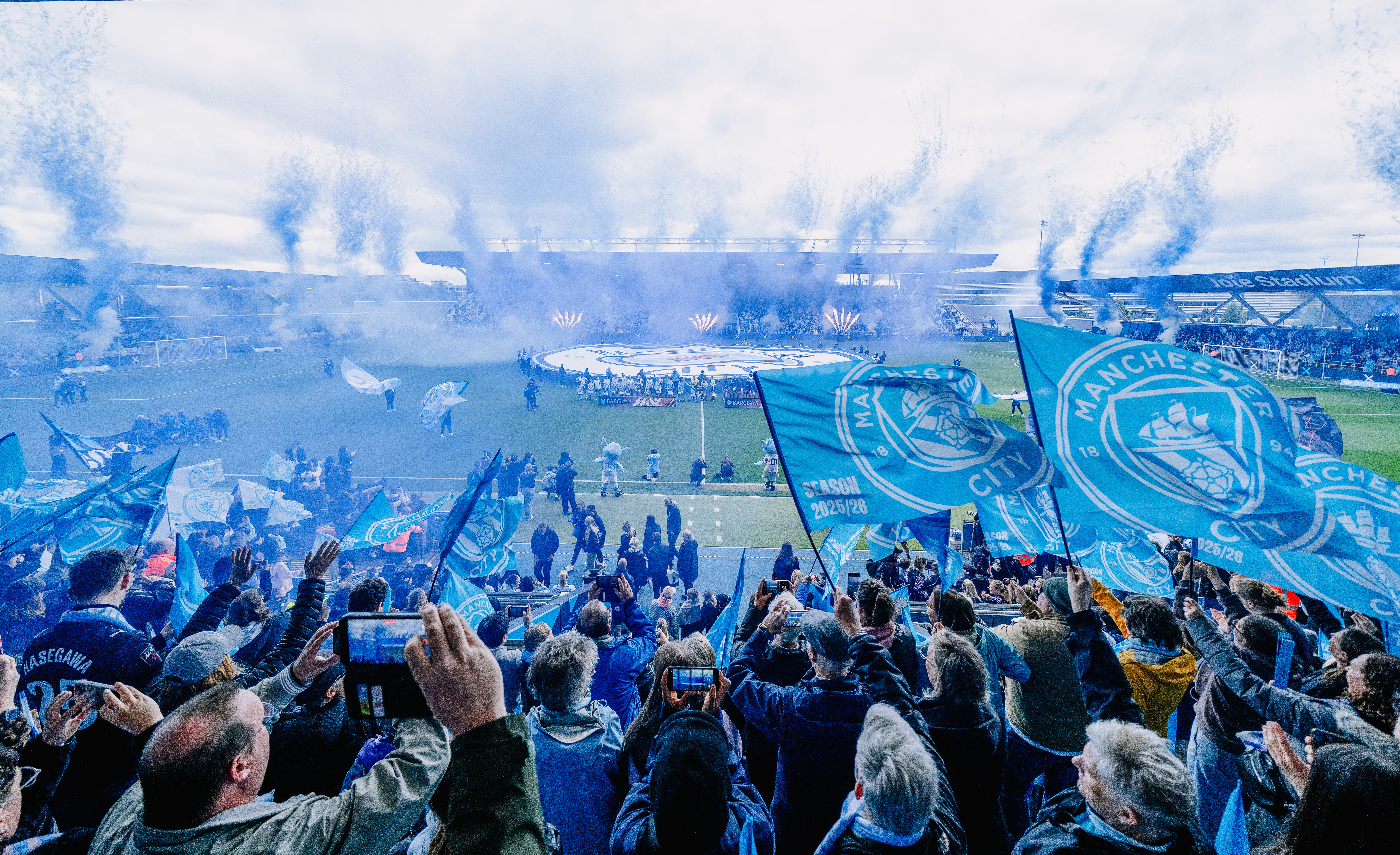 Football fans celebrating at Joie Stadium with Manchester City flags and confetti.