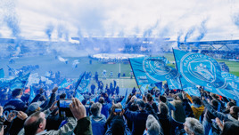 Football fans celebrating at Joie Stadium with Manchester City flags and confetti.