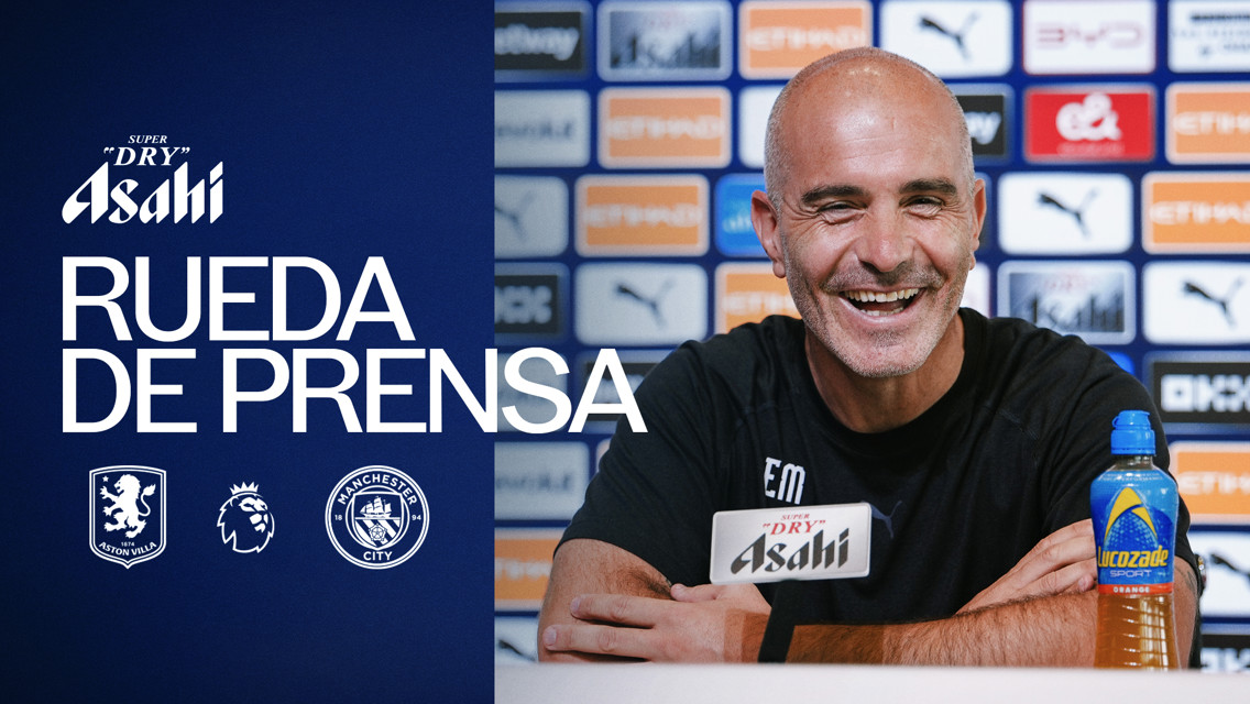 A man smiling at a press conference, sponsored by Asahi, with Aston Villa and Manchester City logos. The backdrop shows various sponsor logos including Puma and Ethihad.