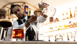 A bartender skillfully pouring a cocktail into a martini glass using a strainer with other drinks on the bar counter.