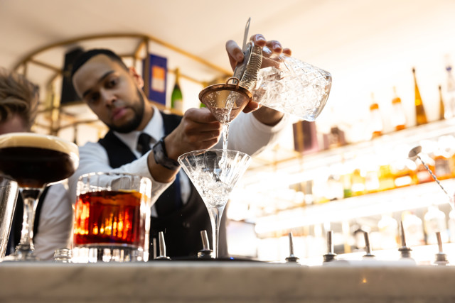 A bartender skillfully pouring a cocktail into a martini glass using a strainer with other drinks on the bar counter.