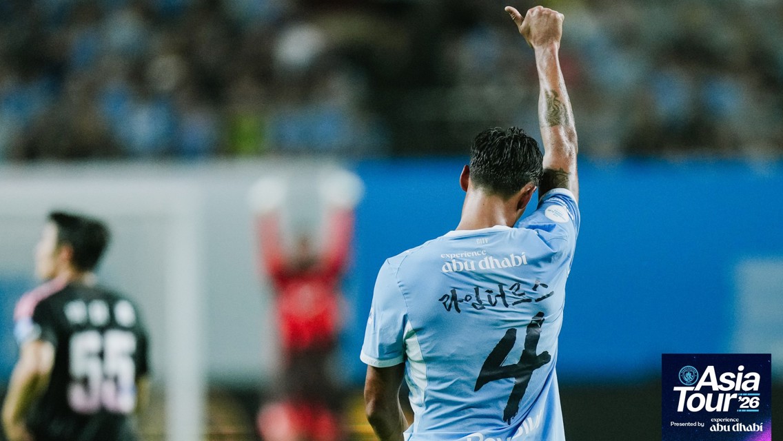 Football player in a light blue jersey with number 4 raises a finger. The shirt has 'experience abu dhabi' text. Backdrop of an indoor stadium. Asia Tour '26 logo in the corner.