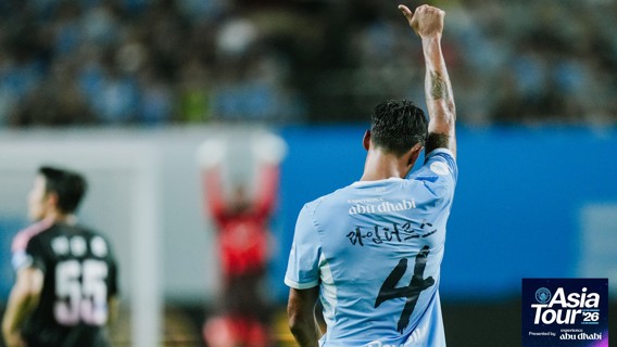 Football player in a light blue jersey with number 4 raises a finger. The shirt has 'experience abu dhabi' text. Backdrop of an indoor stadium. Asia Tour '26 logo in the corner.