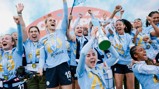 A group of Manchester City Women's football players celebrate holding a trophy after winning a championship, wearing blue jerseys with the number 26 and logos displayed.