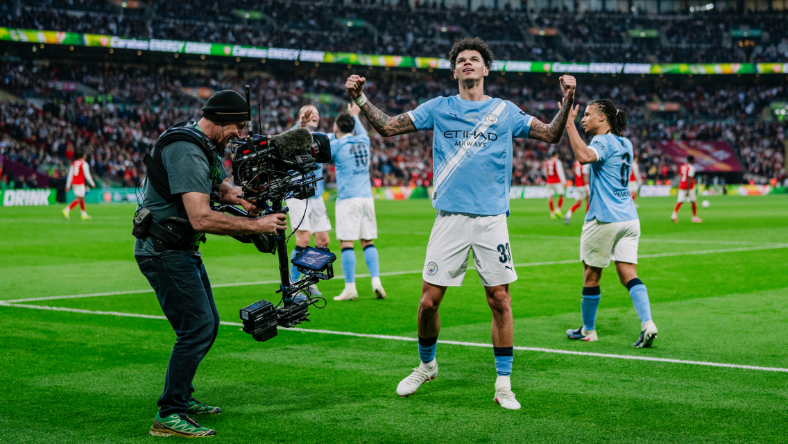 Manchester City players celebrate a goal on the field with a cameraman filming the moment. Players are wearing light blue jerseys, with a large crowd in the background.