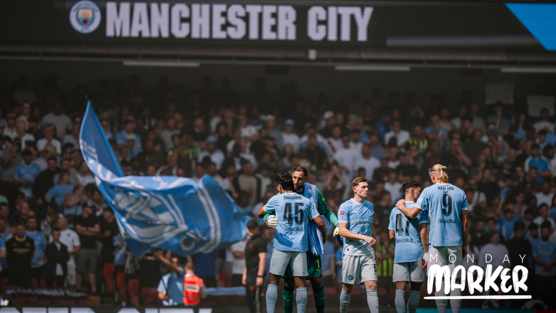 The image shows Manchester City players celebrating on the field, with a large blue Manchester City flag and crowd in the background.