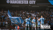 The image shows Manchester City players celebrating on the field, with a large blue Manchester City flag and crowd in the background.