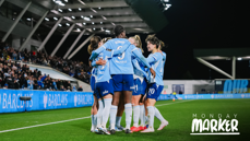 Soccer players wearing light blue jerseys celebrating a goal on the field. The stadium is full with spectators, and 'Monday Marker' text is visible.