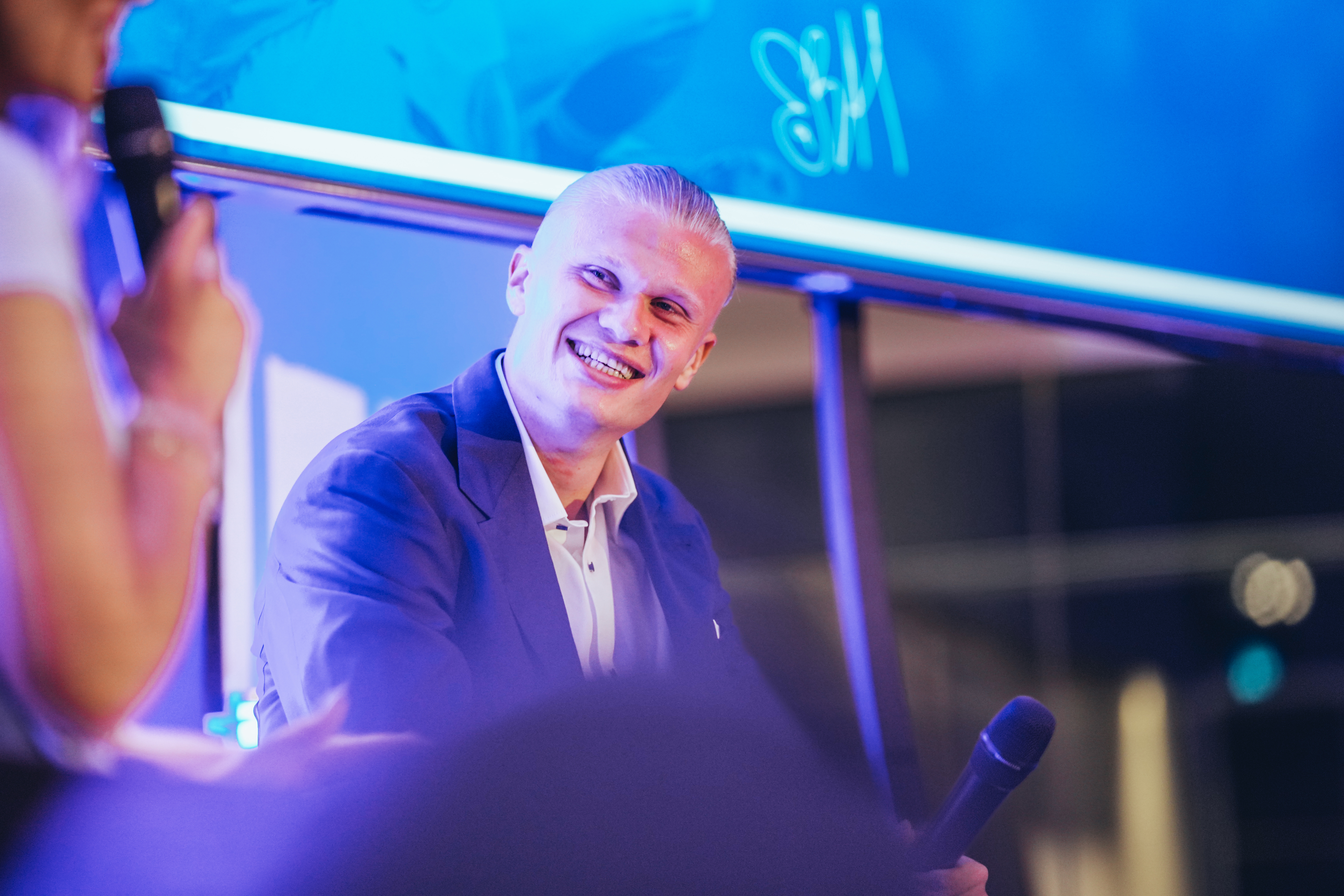 A panel discussion scene with two individuals holding microphones. The face of one person is blurred and they are in front of a large screen with a blue background.
