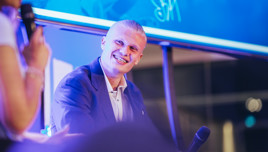 A panel discussion scene with two individuals holding microphones. The face of one person is blurred and they are in front of a large screen with a blue background.