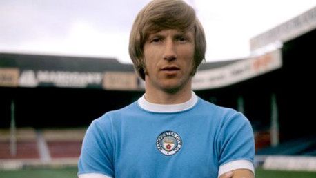 A footballer wearing a vintage Manchester City jersey, standing with arms crossed in a stadium.