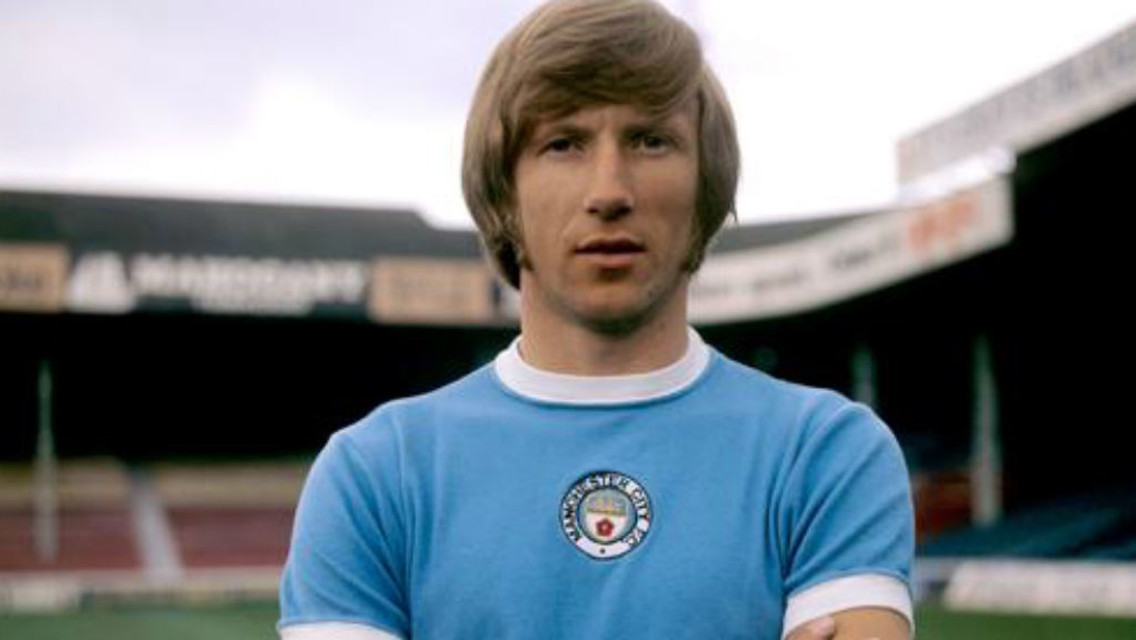 A footballer wearing a vintage Manchester City jersey, standing with arms crossed in a stadium.