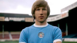A footballer wearing a vintage Manchester City jersey, standing with arms crossed in a stadium.
