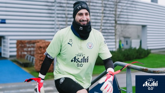 A Manchester City player dressed in training gear, featuring sponsor logos, steps outdoors at the training facility with gloves and knee pads.