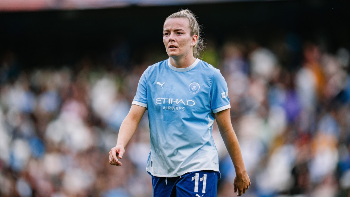 A Manchester City female player wearing the team's sky blue kit with the number 11 visible, walking on the field during a match.