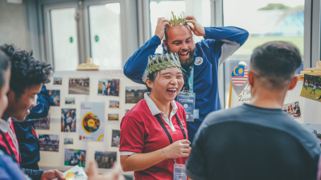 A group of people wearing event badges and crowns made of leaves engage in conversation at an indoor event booth displaying various photos and a Malaysian flag.