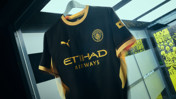 Manchester City black and gold jersey featuring team crest, Puma logo, and Etihad Airways sponsor, hanging in a showcase. Background contains posters with the words Hive state of mind.