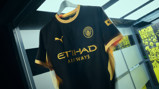 Manchester City black and gold jersey featuring team crest, Puma logo, and Etihad Airways sponsor, hanging in a showcase. Background contains posters with the words Hive state of mind.