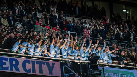 A football team in light blue jerseys celebrates with a trophy in a stadium packed with cheering fans.