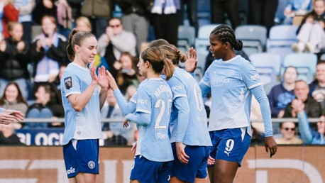 Manchester City women's team players in blue jerseys celebrate a goal with high-fives on the field during a match.
