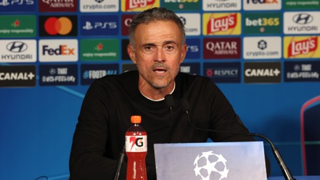A man is speaking at a press conference with a UEFA Champions League backdrop and various sponsor logos. A Gatorade bottle is on the table in front of him.