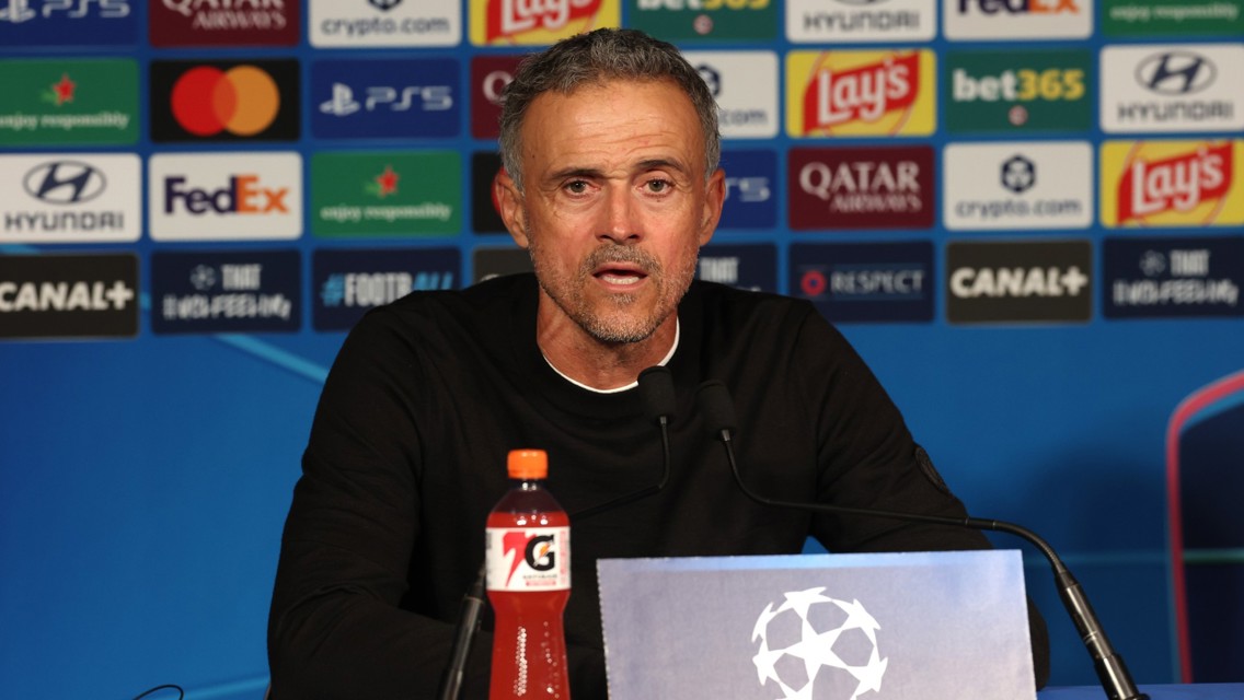 A man is speaking at a press conference with a UEFA Champions League backdrop and various sponsor logos. A Gatorade bottle is on the table in front of him.