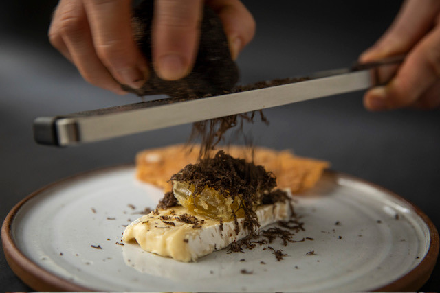A person grates black truffle over a slice of cheese, with bread in the background, showcasing a culinary preparation.