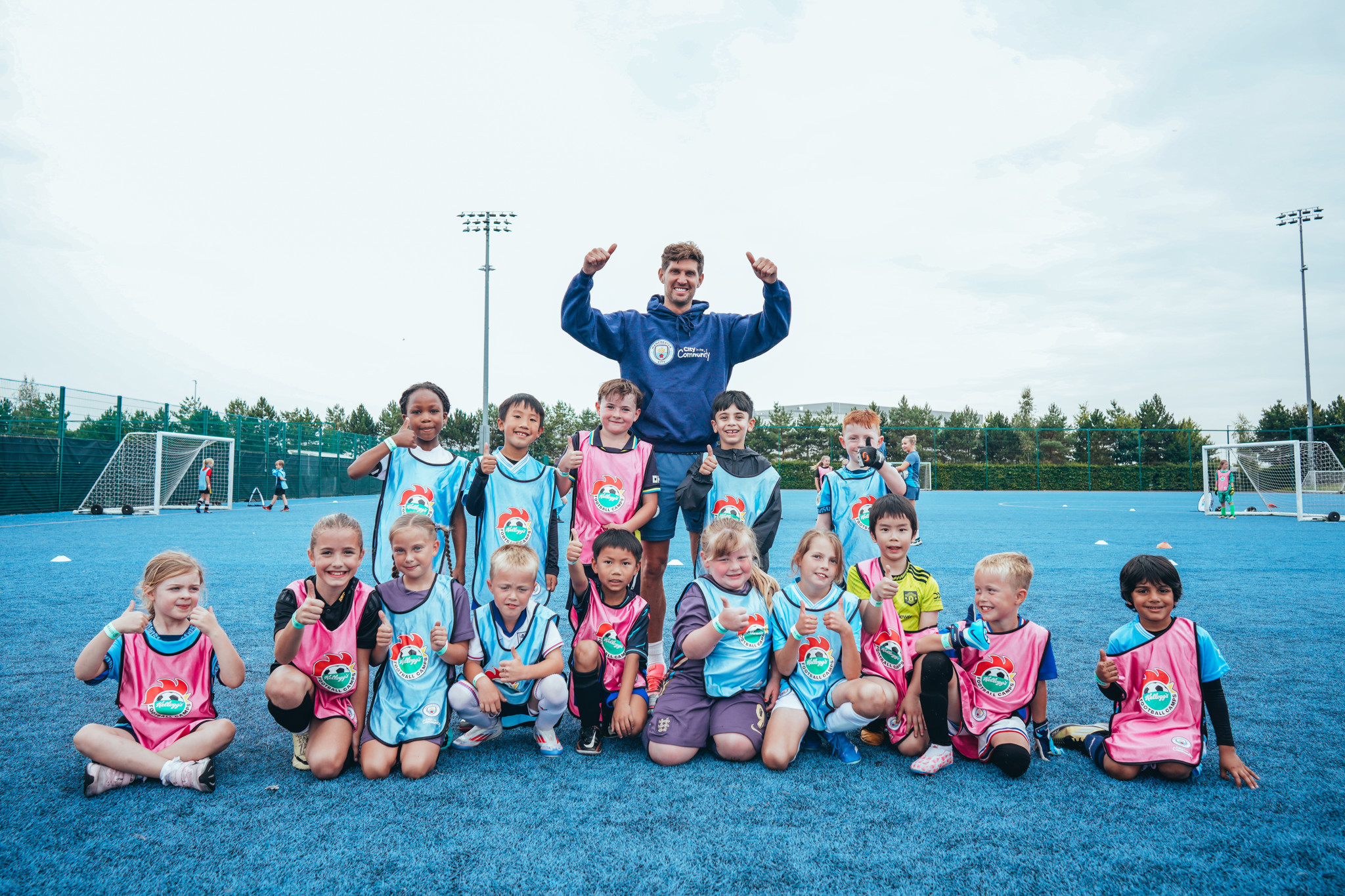 Group photo of children in football kits with a coach on a blue football pitch. Kids are wearing Manchester City community bibs.