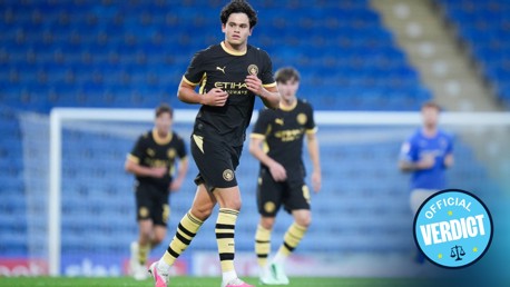 A young football player in a Manchester City kit playing on a field with a blue stadium backdrop and other players in the background wearing similar kits.