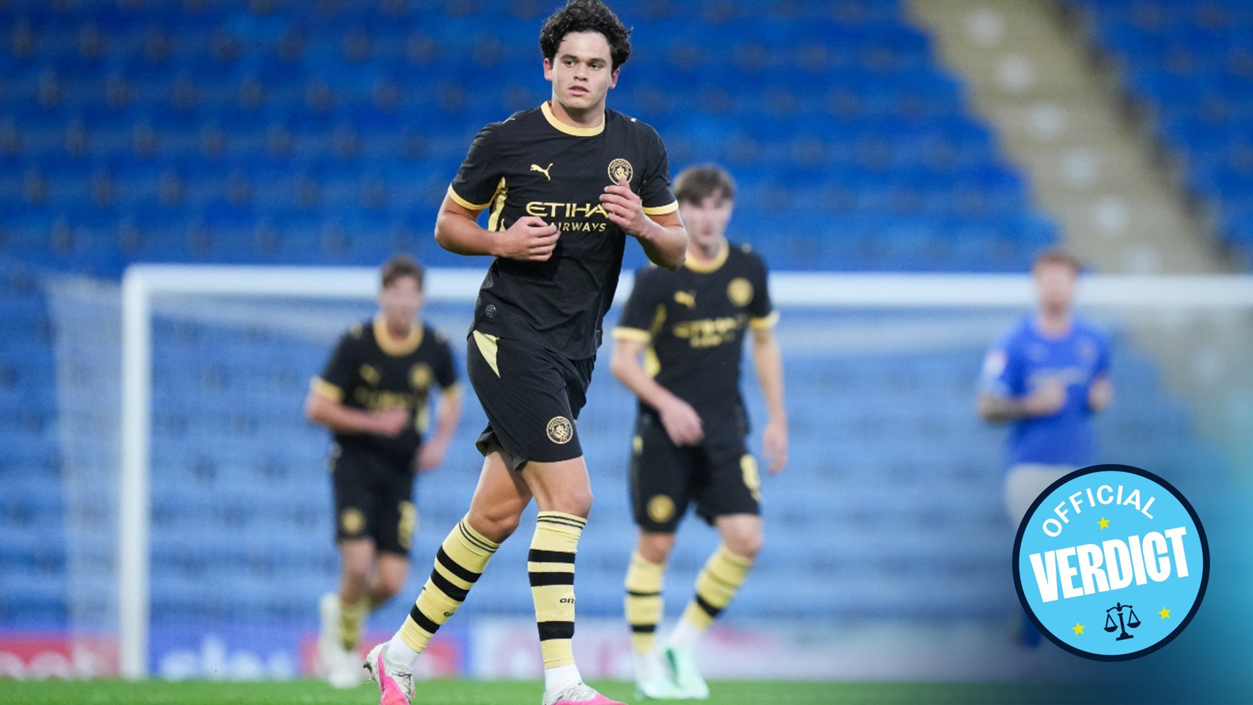 A young football player in a Manchester City kit playing on a field with a blue stadium backdrop and other players in the background wearing similar kits.