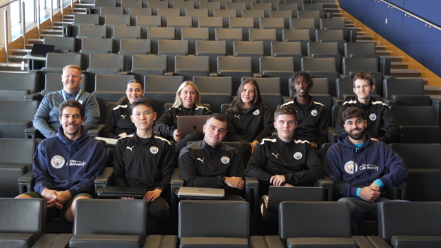 Group of people in Manchester City training jackets in a lecture hall, some holding laptops and notebooks, likely from City in the Community program.