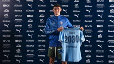 A person in a Manchester City tracksuit holding a jersey with the name 'Foden' and '2030', standing against a backdrop of sponsor logos.