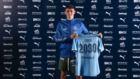 A person in a Manchester City tracksuit holding a jersey with the name 'Foden' and '2030', standing against a backdrop of sponsor logos.
