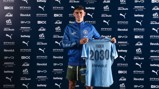 A person in a Manchester City tracksuit holding a jersey with the name 'Foden' and '2030', standing against a backdrop of sponsor logos.