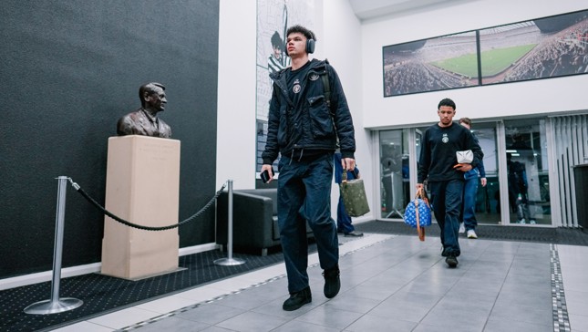 People in a hallway walking past a bust on a pedestal, framed photos of a stadium on the walls.