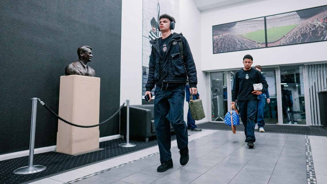 People in a hallway walking past a bust on a pedestal, framed photos of a stadium on the walls.