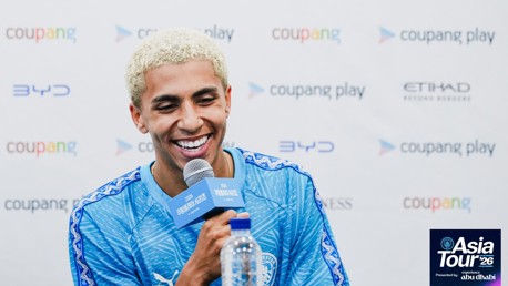 A person with blond hair smiling while holding a microphone at a press conference, wearing a blue Manchester City jersey. The background displays logos including 'Coupang Play' and 'Etihad'. The event is related to the 'Asia Tour '26'.