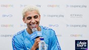 A person with blond hair smiling while holding a microphone at a press conference, wearing a blue Manchester City jersey. The background displays logos including 'Coupang Play' and 'Etihad'. The event is related to the 'Asia Tour '26'.