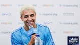 A person with blond hair smiling while holding a microphone at a press conference, wearing a blue Manchester City jersey. The background displays logos including 'Coupang Play' and 'Etihad'. The event is related to the 'Asia Tour '26'.