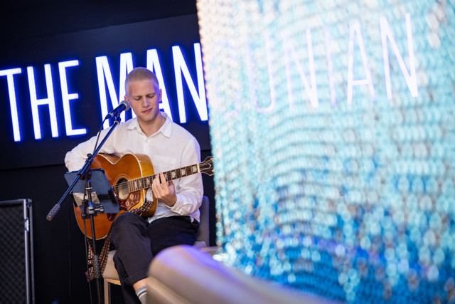 A musician seated, playing an acoustic guitar with a microphone and music stand near him. The background features bright neon text saying 'THE MANCUNIAN'.