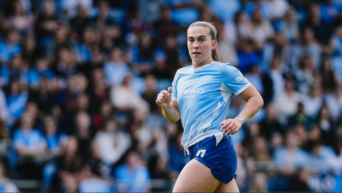 A female Manchester City player in action during a match. She is wearing a light blue kit with the team's logo visible on the jersey. The crowd is blurred in the background.