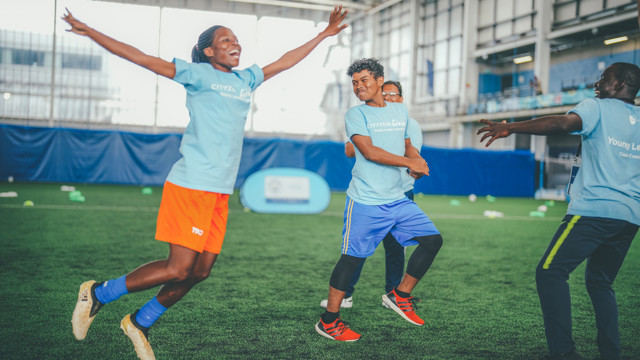 A group of young athletes engaging in a sports training session indoor on a synthetic grass field. The participants wear blue shirts with 'Cityzens Giving' text visible, and one person is jumping energetically.